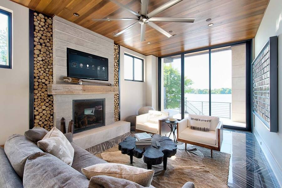 Modern living room with a cozy fireplace framed by stacked firewood, a wall-mounted smart TV, a wooden ceiling with a ceiling fan, and large windows offering a stunning view of a lake.