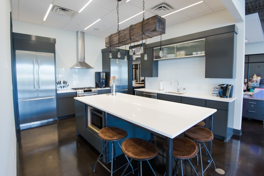 Modern kitchen with dark cabinets, a white island countertop, industrial-style stools, and a rustic wood beam light fixture hanging above.