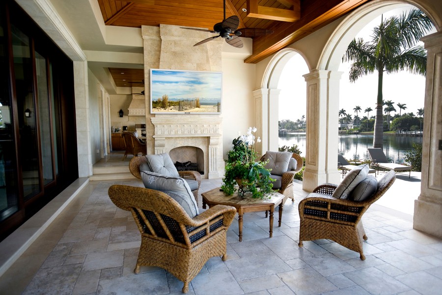 Covered outdoor patio with wicker seating, a fireplace, wall-mounted TV, and tropical lakefront views framed by stone arches and palm trees.