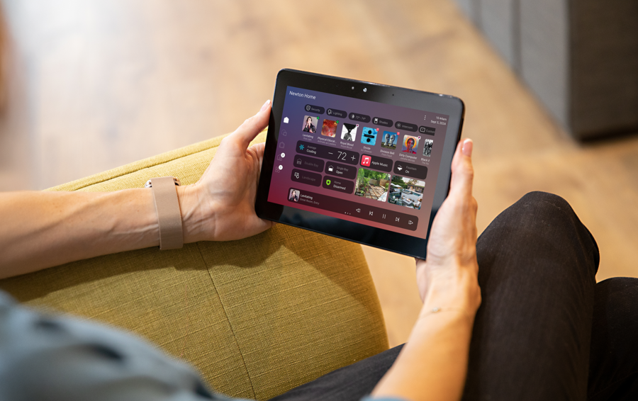 A person holding a tablet displaying a smart home control interface, with options for media, lighting, climate, and home systems, while seated comfortably indoors.